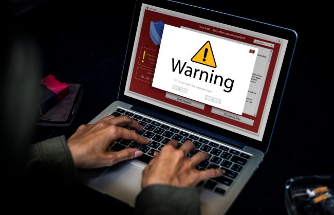 Photo of man typing on laptop with warning sign illustrates blog: "5 Cybersecurity Basics for Small Businesses"